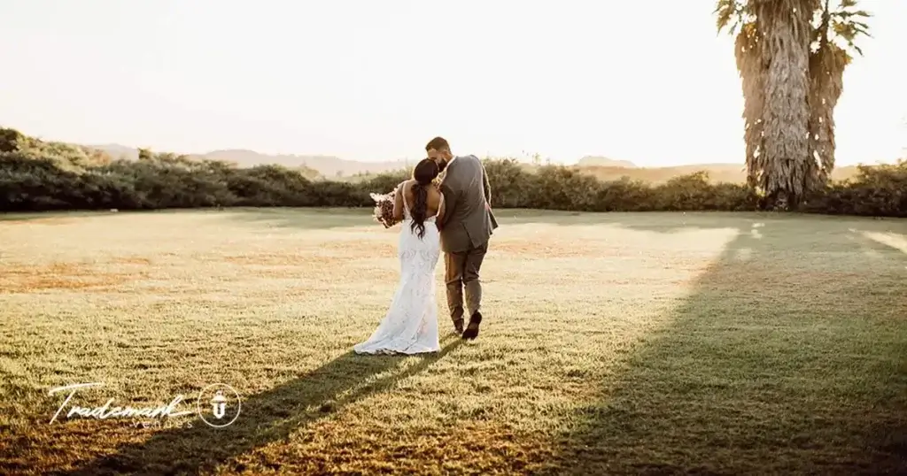 Bride and groom holding hands while standing on a grassy ridge, looking out over a sweeping mountain horizon at a Southern California wedding venue.